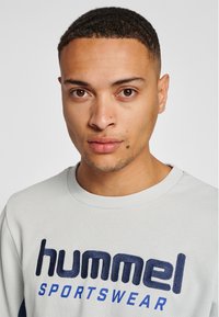 Jasnoszara bluza z niebieskim logo "hummel", okrągły dekolt i ściągacze na rękawach. Miękka w dotyku, casualowy design odpowiedni na co dzień.