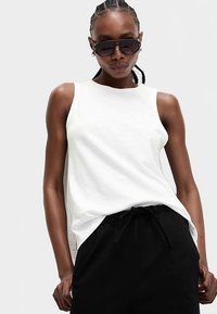 White sleeveless top made of cotton, featuring a crew neckline and a relaxed fit, paired with black drawstring pants.