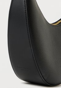 Svart läderhandväska med en slät textur, rund form och en subtil präglad logga. Har guldaccenter och kontrasterande sömmar.