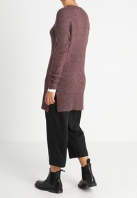 Maglione oversized color bordeaux a maglia con maniche lunghe, polsi a costine e aperture laterali. Indossato sopra pantaloni cropped neri e stivali neri alla caviglia.