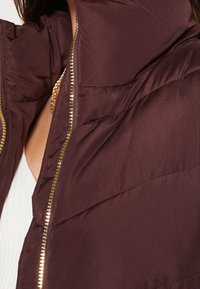 Close-up van een persoon die een donkerbruine gewatteerde jas met een gouden ritssluiting, een wit shirt en een gouden ketting draagt.