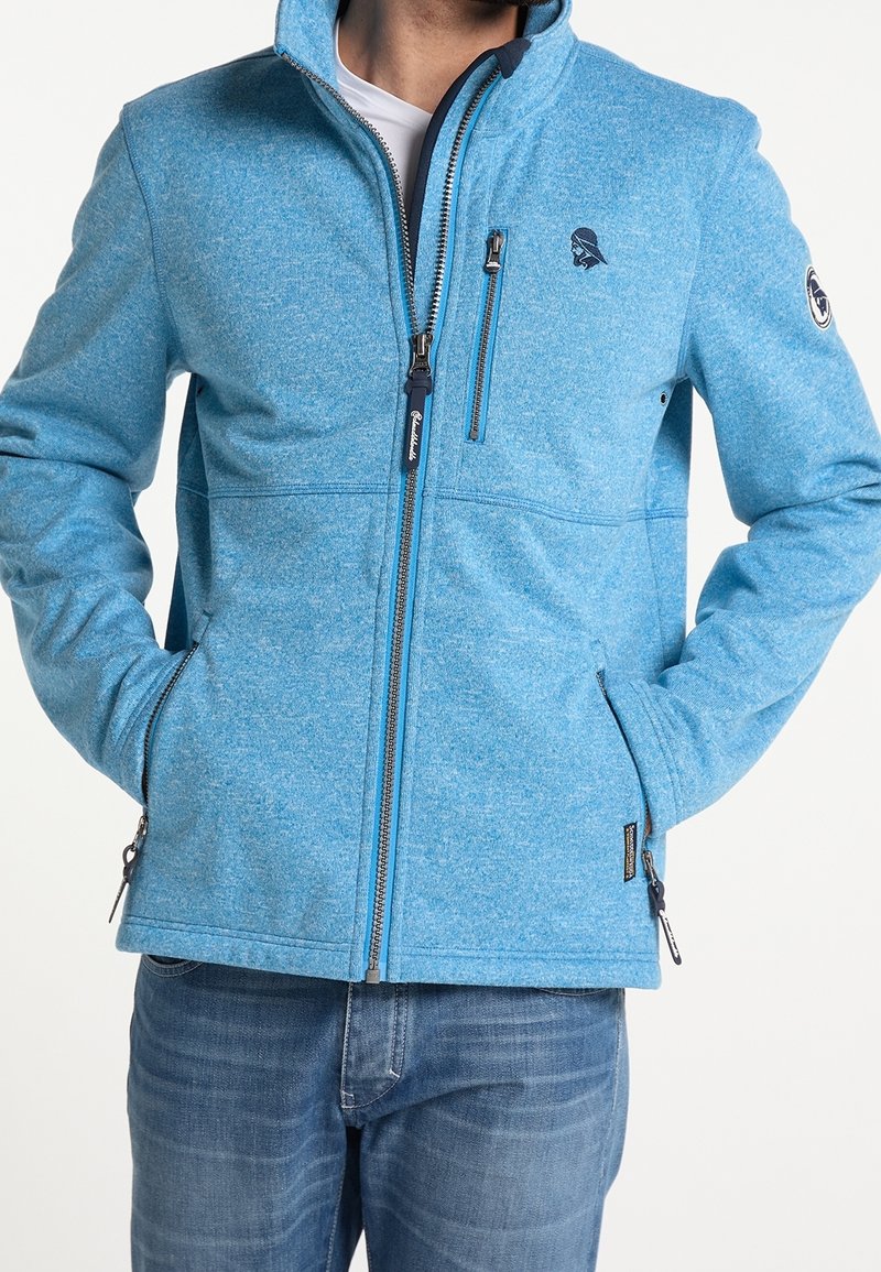 Giacca blu con zip, realizzata in materiale testurizzato, con colletto rialzato e tasche con zip. Presenta il logo sul petto e polsini regolabili. Indossata sopra una camicia bianca.