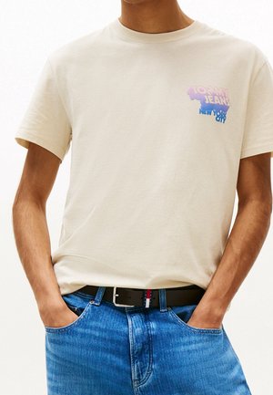 Beige katoenen t-shirt met een geprint logo in paars en blauw, gecombineerd met blauwe jeans en een zwarte riem met een klein rood, wit en blauw accent.