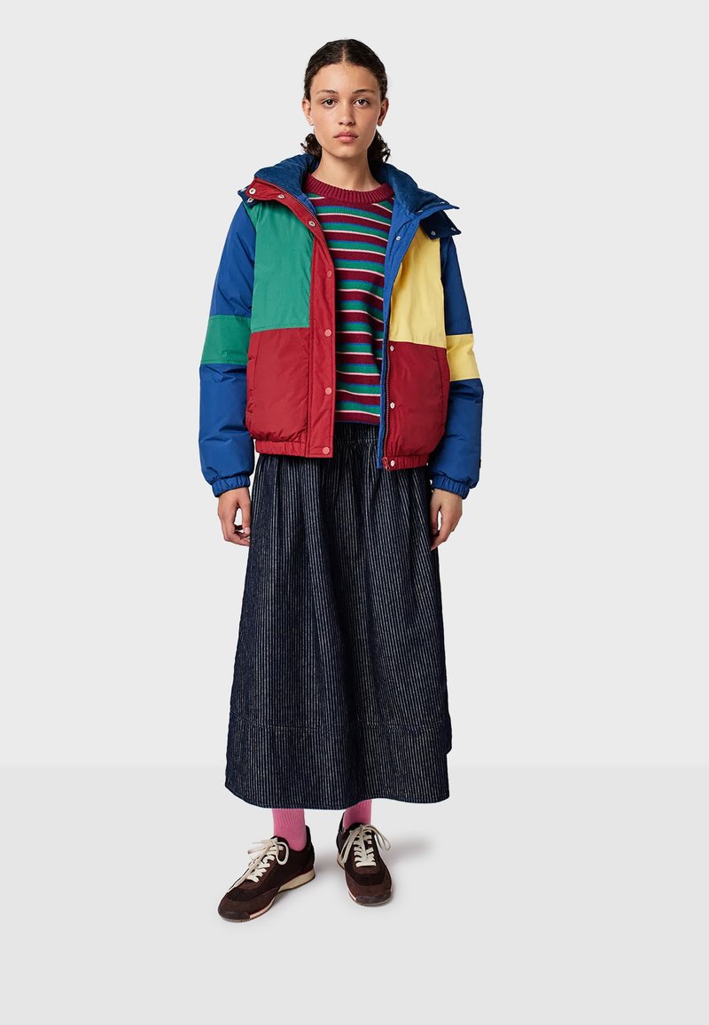 Young woman wearing a multicolored puffer jacket, striped sweater, long dark pleated skirt, pink socks, and brown sneakers standing on a plain background.