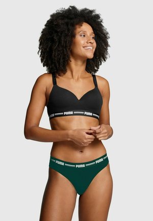 Woman with curly hair wearing black Puma sports bra and dark green Puma briefs, smiling and looking to the side against a plain background.