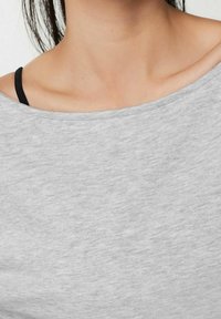 T-shirt en coton doux, gris clair, avec un décolleté large. Les bretelles noires d'un vêtement en dessous sont visibles sur l'épaule gauche.