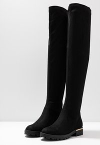 Botas negras de ante por encima de la rodilla, con suela de goma gruesa y un sutil acento dorado en el tacón. Textura suave, diseño aerodinámico.