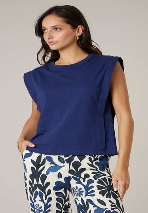 Vrouw die een mouwloos marineblauw topje en witte broek met een blauw-zwart abstract bloemenpatroon draagt, staand met één hand in haar zak.