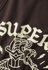 Top de algodón marrón sin mangas con letras agrietadas en un beige claro y un diseño gráfico detallado de una criatura con contornos y acentos en negrita.