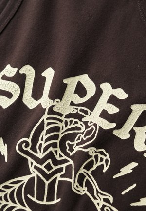 Brązowy bawełniany top na ramiączkach z popękanym jasnobeżowym napisem oraz szczegółowym projektem graficznym stworzenia z wyraźnymi konturami i akcentami.