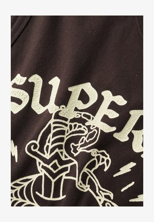 Brązowy bawełniany top na ramiączkach z popękanym jasnobeżowym napisem oraz szczegółowym projektem graficznym stworzenia z wyraźnymi konturami i akcentami.