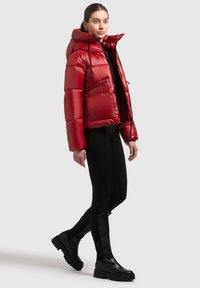 Chaqueta acolchada roja con cuello alto, bolsillos con cremallera y textura brillante, combinada con leggings negros ajustados y botas negras hasta la rodilla.