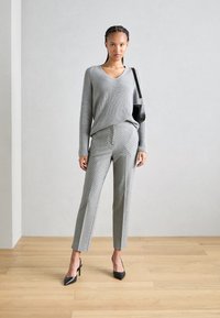Pull en maille gris à col en V, associé à un pantalon à carreaux gris. Des escarpins noirs à bout pointu et un petit sac à main complètent la tenue. Fond en bois.
