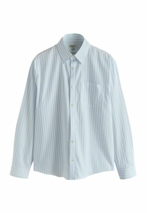 Jeune homme portant une chemise à boutons rayée verticalement bleu clair et blanche, avec un col pointu et une poche poitrine, sur un fond neutre.