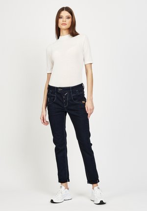 Dünne dunkelblaue Jeans mit Fronttaschen und Knopfverschluss, kombiniert mit einem figurbetonten weißen Kurzarmoberteil und weißen Sneakers.