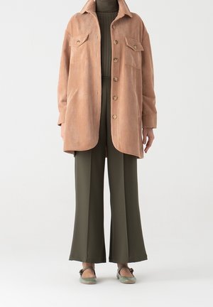 Personne portant une surchemise en velours côtelé beige boutonnée, un pantalon large vert olive, un col roulé assorti et des chaussures mary jane vertes.