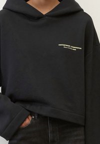 Černá mikina s kapucí vyrobená z měkké látky, se žlutým logem na předním hrudníku. Má volný střih a design s pokleslým ramenem.