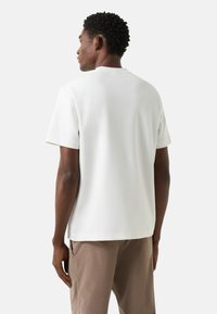 Biała, jednokolorowa koszulka z krótkim rękawem i okrągłym dekoltem, o luźnym kroju i gładkiej fakturze, zestawiona z brązowymi spodniami.