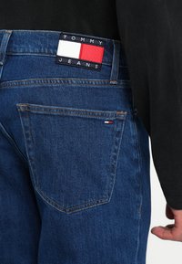 Zilas džinsa bikses ar taisnstūrveida Tommy Jeans logotipa plāksteri, kurā ir sarkanas, baltas un zilas krāsas. Redzama aizmugurējā kabata ar oranžām šuvēm.