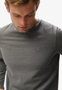 Graues Langarmshirt mit rundem Halsausschnitt, versehen mit einem dezenten, auf die Brust gestickten Logo, aus einem glatten, leichten Stoff gefertigt.