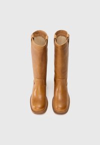 Bottes en cuir marron, hautes et élégantes, avec une texture lisse, un bout arrondi et des fentes sur les côtés en haut. Des coutures discrètes soulignent la semelle.