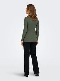 Žena stojaca vľavo, oblečená v olivovo zelenom tričku s dlhým rukávom a čiernych nohaviciach na jednoduchom svetlom pozadí.