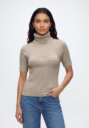 Femme aux longs cheveux foncés portant un pull beige à manches courtes et col roulé, ainsi qu’un jean bleu, debout avec une main dans la poche sur fond blanc.