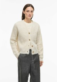 Kremasto obarvan kardigan iz mehkega, puhastega materiala z izrezom v obliki črke V, petimi okroglimi gumbi in rahlo skrajšanim robom.