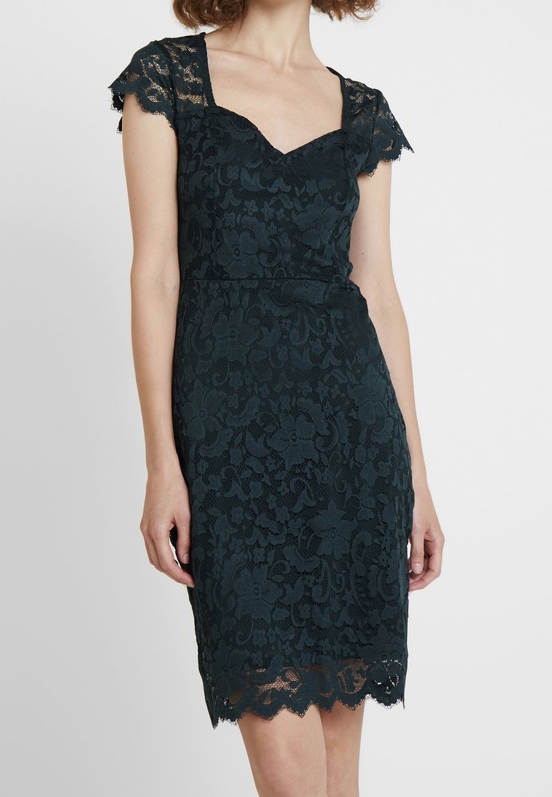 Tmavě tyrkysové krajkové šaty s přiléhavým střihem, krátkými vroubkovanými rukávy a květinovým vzorem; vroubkovaný lem přidává dekorativní detail.