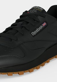 Baskets Reebok Classic noires avec lacets noirs, semelle en gomme et logo Union Jack sur le côté près du nom de la marque.