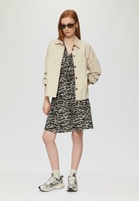 Beige corduroy jacket with wooden buttons worn over a black and white abstract patterned dress. The outfit is completed with silver sneakers and light gray socks.