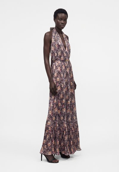 Γυναίκα που φοράει αμάνικο μακρύ φόρεμα με floral σχέδιο και λαιμόκοψη halter, με βολάν στο τελείωμα και μαύρα σανδάλια με ψηλό τακούνι, στέκεται μπροστά σε απλό φόντο.