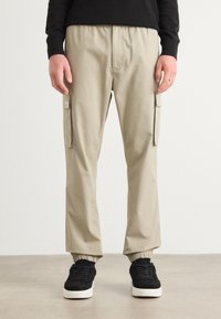 Beige cargo-housut, joissa on joustava vyötärö, sivutaskut ja ribatut hihnat, yhdistetty mustiin lenkkareihin, joissa on valkoinen pohja.