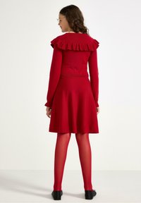 Nainen, jolla on polvipituinen punainen mekko, jossa on röyhelökaulus, punaiset legginsit, mustat ballerinat ja silmälasit, seisoo selin valkoista taustaa vasten.