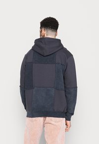 Felpa con cappuccio in patchwork blu navy, dal texture morbido, con cappuccio e vestibilità ampia. Pannelli a contrasto sul retro offrono un interesse visivo.