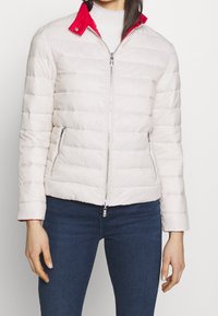 Lehká péřová bunda v krémové barvě s jasně červeným límcem. Má zapínání na zip, boční kapsy a prošívanou strukturu.