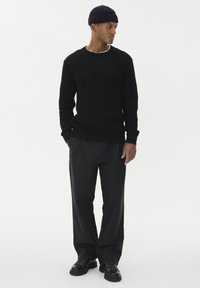 Man som bär en svart stickad tröja, svarta byxor, en svart mössa och svarta snörskor, står med händerna i fickorna och ser åt höger.