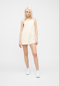 Νεαρή γυναίκα με μακριά ξανθά μαλλιά, φορώντας ένα αμάνικο κρεμ ολόσωμο σορτς με υφή και λευκά αθλητικά παπούτσια, στέκεται σε απλό λευκό φόντο.