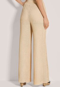 Beige strukturierte weite Hose, getragen mit Plateauschuhen aus Geflecht, von hinten gezeigt, mit einer Hand auf der Hüfte. 