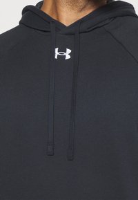 Gros plan sur une personne portant un sweat à capuche noir avec un logo Under Armour blanc sur la poitrine et des cordons noirs.