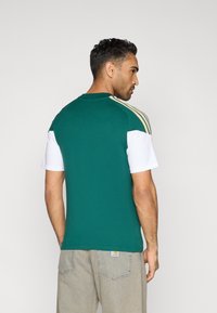 Grøn og hvid kortærmet skjorte med rund hals, tekstureret stof og tre horisontale striber på skuldrene i guld.