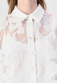Hvid bluse med blomsterbroderi, krave, gennemsigtig stof og knaplukning foran med klare knapper. Teksturede blomster mønstre.