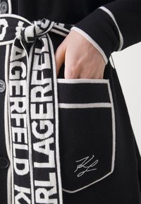Hand in der Tasche einer schwarzen Strickjacke mit weißem Besatz, die einen schwarz-weißen Gürtel mit dem Text "LAGERFELD" und einer signierten Stickerei aufweist.