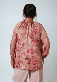 Bluse aus transparentem rosa Stoff mit gerafftem Kragen, langen Ärmeln und lebhaften Blumenmustern in verschiedenen Rosa- und Lilatönen.