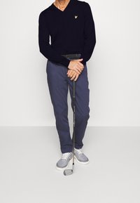 Pull noir à col en V, pantalon bleu marine, chaussures de sport grises. Club de golf tenu dans les mains, avec une poignée texturée et une tête de club argentée.