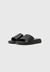 Sandales noires à glissière avec une tige en cuir lisse, un design à bout ouvert et une semelle en caoutchouc plate, présentant une forme minimaliste et moderne.