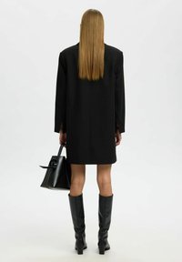 Černé přizpůsobené sako-dress s dlouhými rukávy, spadlými rameny a rovnou siluetou. Modelka drží černou kabelku a nosí kozačky do kolen.