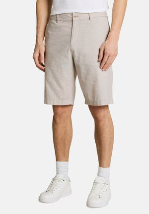 Mann trägt beige knielange Shorts, weiße Sportsocken und weiße Turnschuhe mit Schnürsenkeln, steht vor einem schlichten Hintergrund.