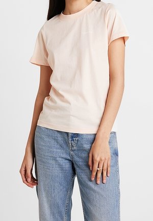 Frau trägt ein hellrosa kurzärmeliges T-Shirt und hellblaue Jeans, steht mit einer Hand an der Hüfte und der anderen locker.