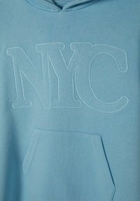 Hanorac de culoare albastru deschis, cu un buzunar tip kangaroo în față și un text mare brodat "NYC" în fir de culoare albastru ușor mai închis. Textura materialului este moale.